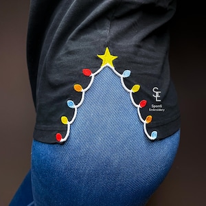 May include: Black long-sleeve shirt with a Christmas tree design embroidered on the side. The tree is made of colorful lights and a yellow star. The shirt is paired with blue jeans. The text "Sponti Embroidery" is visible.