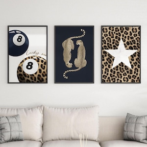 May include: Three framed wall art prints. One features a black and white 8-ball, another shows two cheetahs, and the third has a white star on a leopard print background. The prints are displayed above a gray sofa.