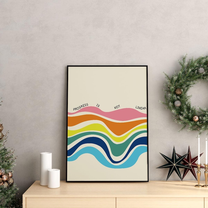 May include: A framed art print with the text "PROGRESS IS NOT LINEAR" above colorful, wavy lines. The artwork features shades of pink, orange, yellow, green, and blue. The print is displayed on a wooden surface with holiday decorations.