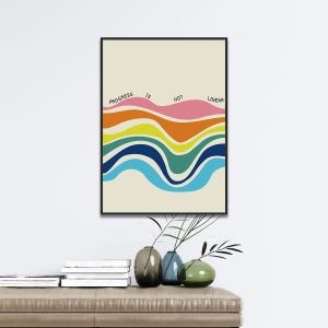 May include: A framed art print with the text "PROGRESS IS NOT LINEAR" above colorful, wavy lines. The artwork features a cream-colored background with bands of pink, orange, yellow, green, blue, and light blue. The frame is black.