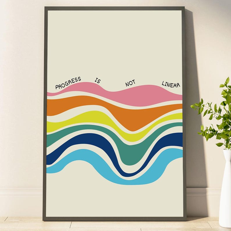 May include: A framed art print with the text "PROGRESS IS NOT LINEAR" above a series of colorful, wavy horizontal bands. The bands include shades of pink, orange, yellow, blue, and teal against a cream background.