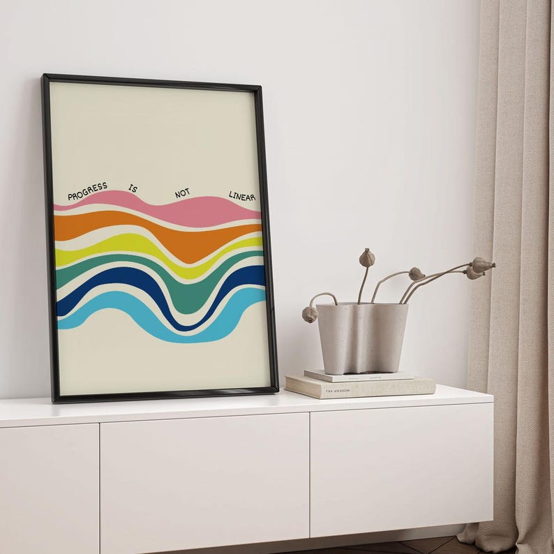 May include: A framed art print with the words "PROGRESS IS NOT LINEAR" above a series of colorful, wavy lines. The waves are in shades of blue, green, yellow, orange, and pink. The artwork is in a black frame and is displayed on a white cabinet.