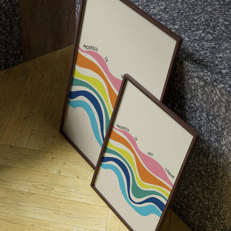 May include: Two framed art prints with a cream background and a colorful, wavy design. The text "Progress is not linear" is printed on the artwork. The frames are a dark brown color.