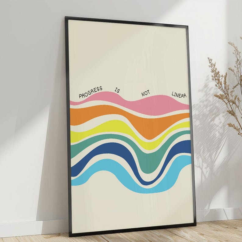 May include: A framed art print with the words "PROGRESS IS NOT LINEAR" above a series of colorful, wavy lines. The lines include shades of pink, orange, yellow, green, and blue against a cream background. The frame is black.