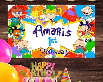 May include: A colorful birthday banner featuring characters from the Nickelodeon cartoon Rugrats. The banner has a yellow starburst with the text "Amari's 1st Birthday" in purple and blue lettering. The banner is decorated with colorful paint splatters.