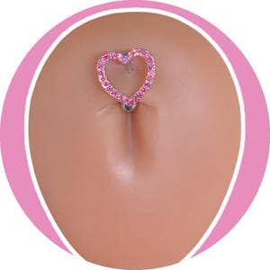 May include: A belly button ring featuring a heart-shaped design. The heart is outlined with small, pink gemstones. The ring is silver-colored and has a small ball at the bottom.