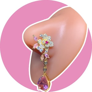 Könnte beinhalten: Ein goldfarbener Nasenring mit floralem Design, verziert mit rosa und klaren Edelsteinen. Ein tropfenförmiger rosa Edelstein hängt von unten. Der Nasenring ist vor einem rosa Hintergrund platziert.