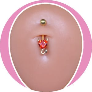 May include: A gold-colored belly button ring with a red heart-shaped gem and devil horn accents. The top ball is gold. The ring is displayed on a light skin tone.