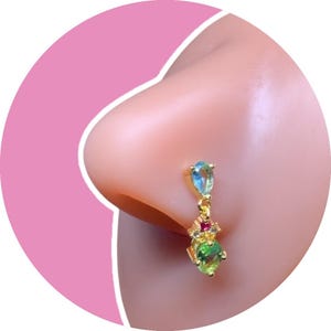 May include: A gold-colored nose ring with a dangling design. The nose ring features a teardrop-shaped light blue gem, a cluster of small red gems, and a round green gem. The jewelry is set against a pink background.