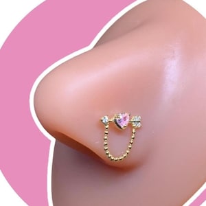 Könnte beinhalten: Ein goldfarbener Nasenring mit einem rosa herzförmigen Edelstein und Pfeildetails. Eine zarte Kette verbindet das Herz mit einem passenden Pfeil. Der Schmuck wird an der Nase einer Person gezeigt.