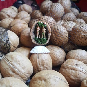 May include: A display of walnuts, some in netting, with one walnut carved to show a miniature scene of two figures in a garden. A seashell decorates another walnut. The backdrop is a colourful, patterned textile.