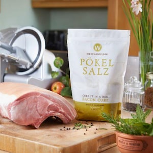 May include: A raw pork roast sits on a wooden cutting board next to a bag of "Pökel Salz" bacon cure. The white bag has gold lettering and a gold bottom. A meat slicer and herbs are in the background.