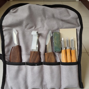 May include: A gray fabric tool roll with a black trim, holding various wood carving tools. The tools include knives with wooden handles and metal blades, along with small chisels and a sharpening stone. The tools are neatly organized in individual pockets.