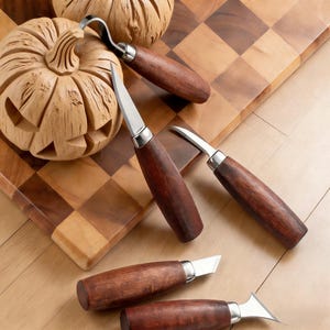 Puede incluir: Un conjunto de herramientas para tallar madera con mangos de madera oscura y hojas plateadas, dispuestas cerca de una calabaza de madera tallada. Las herramientas están diseñadas para trabajos de tallado detallados. El fondo incluye una tabla de cortar de madera y una calabaza tallada.