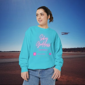 May include: A turquoise blue sweatshirt with the text "Sky Goddess" in pink neon lettering. The sweatshirt also has a graphic of an aeroplane and the text "Currently in" above a blank space.