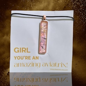 May include: A gold necklace with a rectangular pendant featuring a pink and white map design. The necklace is displayed on a white card with the text "GIRL YOU'RE AN amazing aviatrix!"