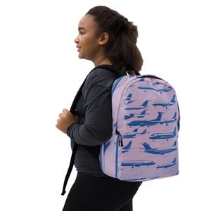 May include: A pink backpack with a blue airplane pattern. The backpack has adjustable straps and a zip closure.