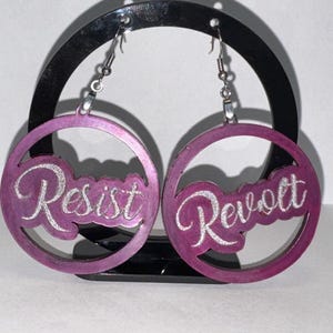 May include: A pair of purple hoop earrings with the words "Resist" and "Revolt" in a cursive font. The earrings are displayed on a black stand against a white background. The text is in a silver glittery color.