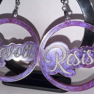 May include: A pair of hoop earrings with a purple and silver design. The word "Revolt" is on one earring and "Resist" on the other. The earrings have a marbled effect and are suspended from silver hooks.