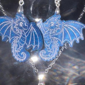 May include: A pair of blue dragon earrings and a matching necklace. The dragon pendants are detailed with white accents and glitter. The jewelry hangs from silver-colored chains and hooks, creating a whimsical and fantasy-themed set.