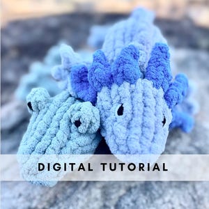 May include: Two crocheted alligator plushies, one blue and one light blue, with the text "DIGITAL TUTORIAL" across the bottom of the image.