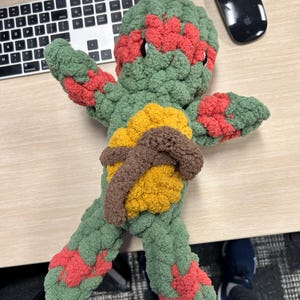 May include: A green, red, and yellow crocheted turtle toy. The turtle has a brown belt around its waist.