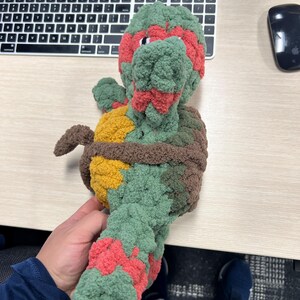 May include: A green, red, brown, and yellow crocheted plush toy shaped like a Teenage Mutant Ninja Turtle. The toy is standing on a wooden surface.