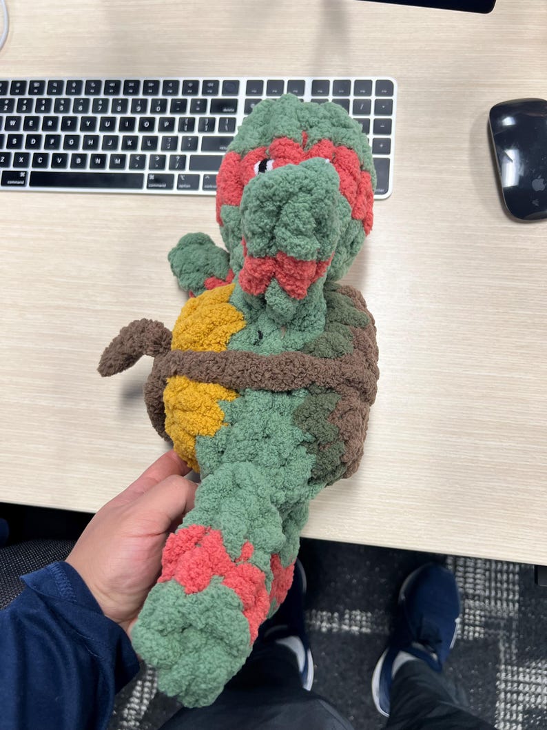 May include: A green, red, brown, and yellow crocheted plush toy shaped like a Teenage Mutant Ninja Turtle. The toy is standing on a wooden surface.