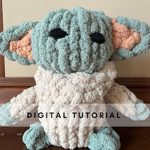 May include: A crocheted plush toy in the shape of a popular fictional character. The toy is green with peach-colored ears and black eyes. The toy is wearing a cream-colored crocheted scarf. The text "DIGITAL TUTORIAL" is visible at the bottom of the image.