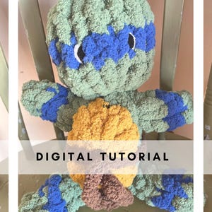 May include: A green, blue, and yellow crocheted stuffed toy turtle with a brown nose. The turtle is wearing a blue eye mask. The text "DIGITAL TUTORIAL" is visible below the turtle.
