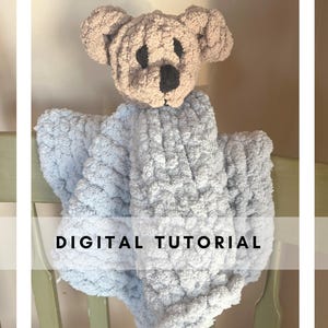 Può includere: Una copertina per neonati in crochet blu chiaro con una testa di orso in crochet marrone attaccata. La copertina è appesa a una sedia di legno. Il testo "DIGITAL TUTORIAL" è stampato sulla copertina.