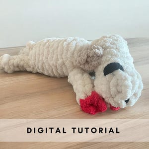 Könnte beinhalten: Ein weißes, gehäkeltes Stofftier in Form eines Hundes mit schwarzen Augen und einem roten Herzen in seinen Pfoten. Das Spielzeug liegt auf einer Holzoberfläche. DIGITAL TUTORIAL steht in schwarzer Schrift auf weißem Hintergrund unter dem Spielzeug.