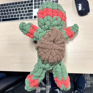 May include: Green and red crocheted turtle plush toy with a brown shell. The toy is lying on a wooden surface.
