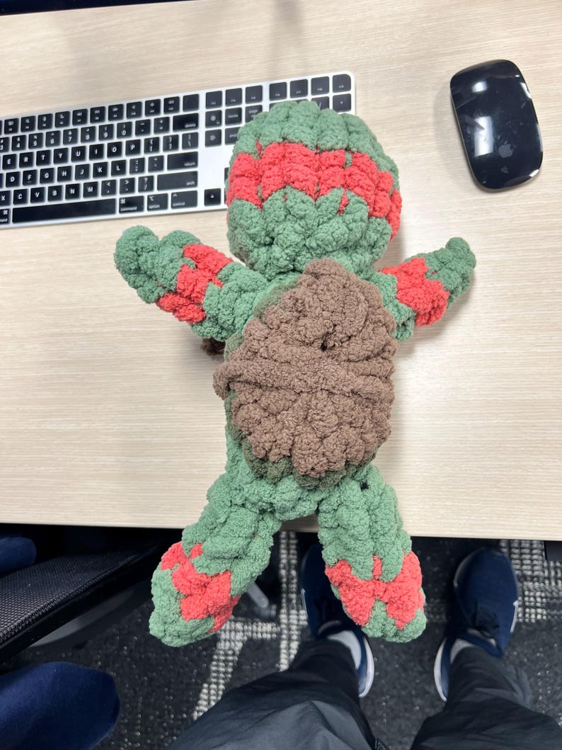 May include: Green and red crocheted turtle plush toy with a brown shell. The toy is lying on a wooden surface.