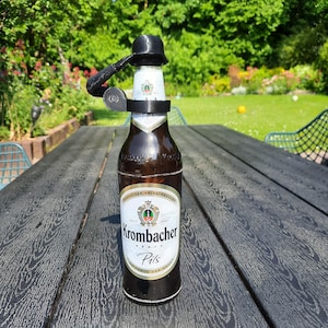 Op de afbeelding: Een bruine glazen bierfles met een zwarte flesopener bevestigd. De fles heeft een wit etiket met de tekst "Krombacher Pils". De flesopener heeft de vorm van een kleine hoed.