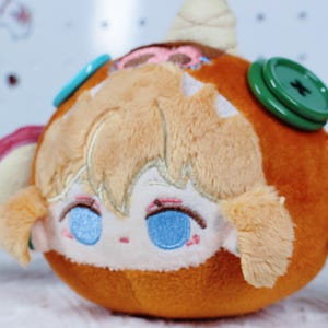 May include: A plush toy with a round, orange body and a detailed face. The face has blue eyes, pink accents, and light brown hair. It features a green button and a light blue button on the side. The toy has a whimsical, cartoonish design.
