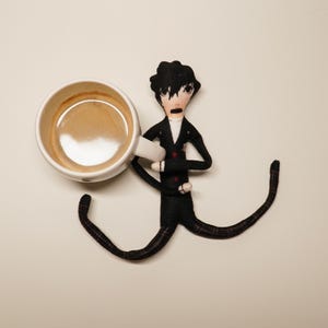 May include: A quirky doll figure in a black suit with red details, holding a white coffee cup filled with coffee. The doll has a surprised expression. The doll's legs are long and patterned.