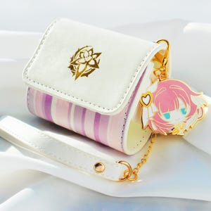May include: A white and striped wallet with a gold-colored charm. The wallet has a white flap with a gold floral design. The body of the wallet is striped with pink and purple. A gold charm of a cartoon character with pink hair and blue eyes hangs from the wallet.