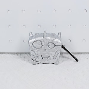 May include: Silver-colored AirPod case with a skull design. The case is square-shaped with a hinged lid and a black carabiner clip attached. The skull features eye sockets, a nose, and a jagged mouth.