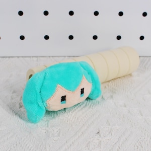 Pode incluir: Um brinquedo de pelúcia com um design de cabelo turquesa e olhos quadrados pretos. O brinquedo está sobre uma superfície branca texturizada, com um objeto cilíndrico bege ao fundo.