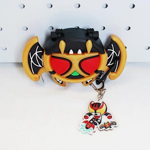 May include: A character-themed keychain in black, gold, and red. The main charm is a stylized bat-like design with red eyes and a green accent. A smaller charm features a cartoon character with a red and yellow colour scheme. The keychain is attached to a silver clasp.