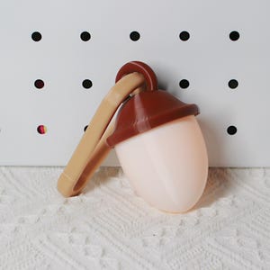 May include: A light brown and dark brown acorn-shaped clip. The clip is attached to a light pink, egg-shaped object with a dark brown cap. The clip is tan and curved. The background is white with black dots.