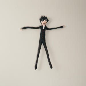May include: A handmade doll in a black suit with red buttons, white shirt, and plaid trousers. The doll has black hair and is posed with arms outstretched. The doll's facial expression is one of surprise or shock.