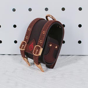 May include: A brown leather mini backpack with gold-colored hardware, including buckles and a ring. The bag features a red emblem and is designed with a small pocket. The stitching and details are visible.