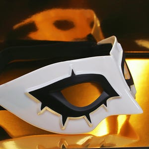 May include: A white and black mask with a stylized design. The mask features a white base with black accents around the eye openings and gold-colored details. The mask is likely for cosplay or costume use.