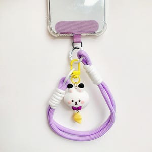 Puede incluir: Un cordón morado para teléfono con un dije de conejo de dibujos animados blanco y amarillo. El cordón tiene una sección blanca y un mosquetón amarillo. El conejo tiene una pajarita morada y una zanahoria amarilla.