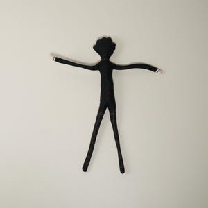 May include: A handmade black fabric doll in a stick figure design, with outstretched arms and plaid patterned legs. The doll has a round head and small white accents on the ends of the arms. A small red heart is sewn on the torso.