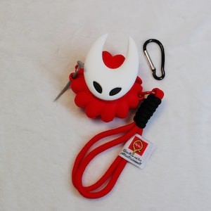 Peut inclure: Un étui pour accessoires rouge et blanc sur le thème de Hornet, avec un mousqueton noir et une lanière rouge. L'étui présente un design de masque blanc avec des yeux noirs et une base rouge en forme de fleur. Une petite étiquette avec du texte est attachée à la lanière.