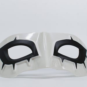 May include: White eye mask with black accents. The mask features black shapes around the eye openings and pointed details. A black strap is attached to the side. The mask is designed for costume or theatrical use.
