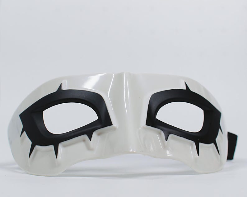 May include: White eye mask with black accents. The mask features black shapes around the eye openings and pointed details. A black strap is attached to the side. The mask is designed for costume or theatrical use.